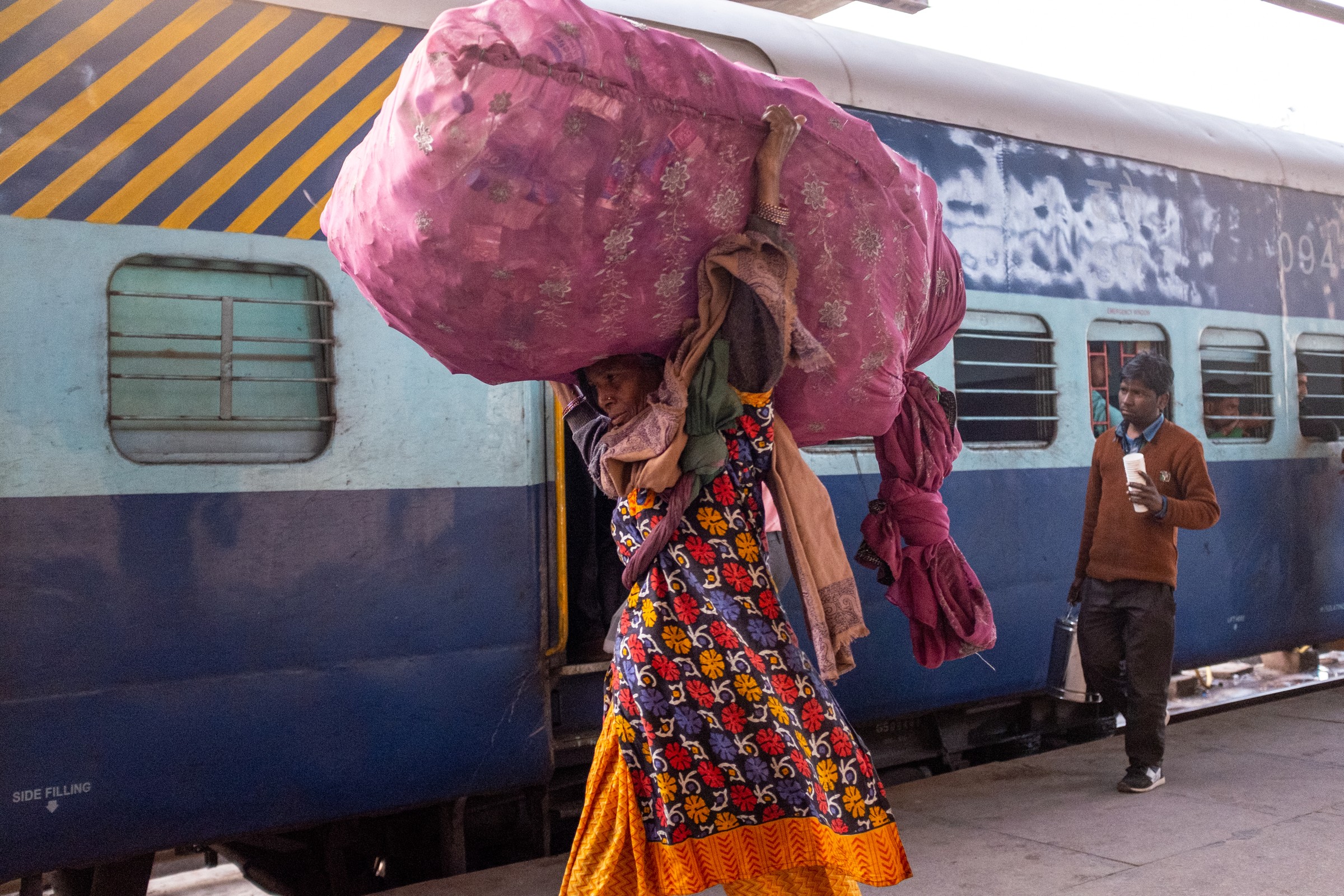 A woman carrying a pink bundle walks beside a blue passenger train.
