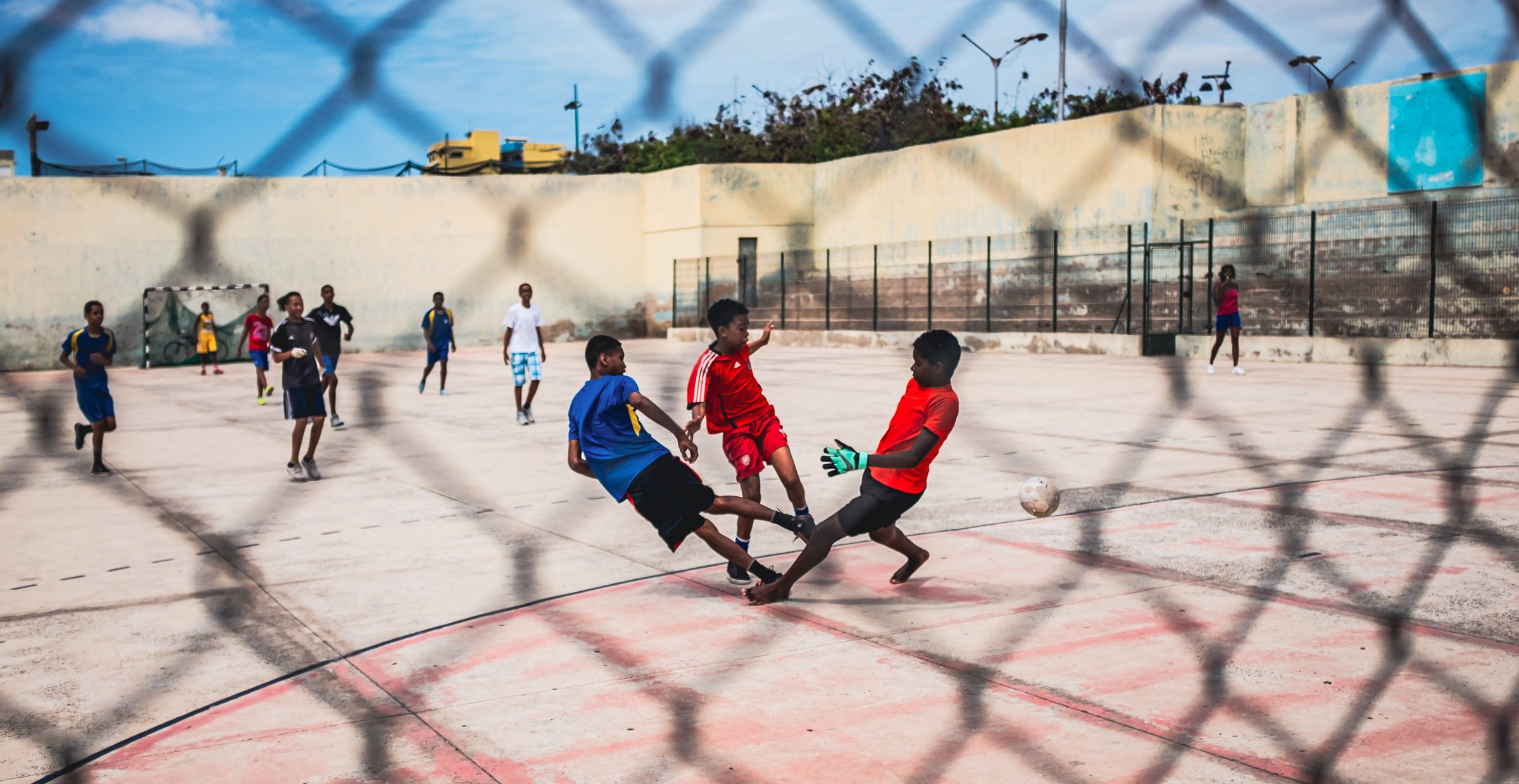 Young players challenge for a football, seen through a wire fence.